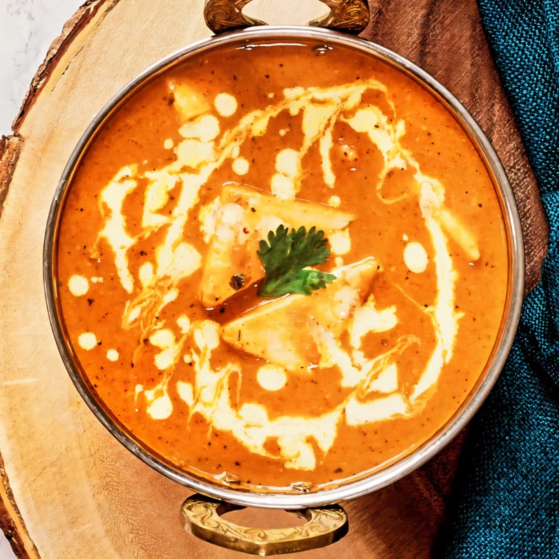 A close-up of Indian paneer butter masala in a metal bowl with cream swirls and cilantro.